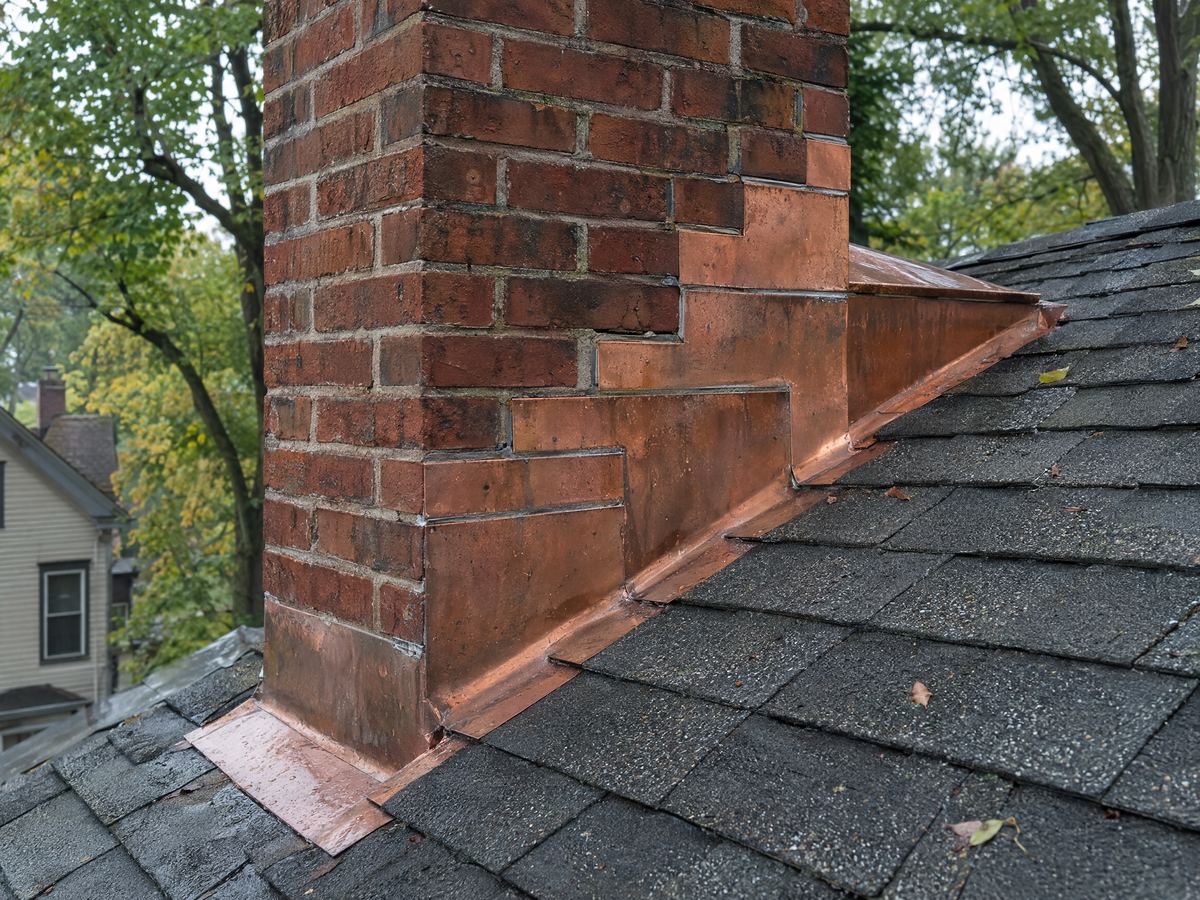 Copper step and counter-flashing where a brick chimney meets a wet asphalt-shingle roof, a copper cricket behind it